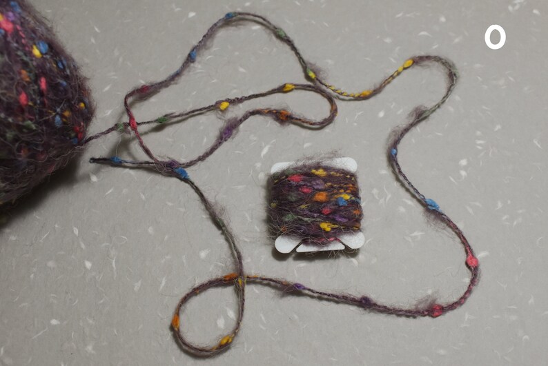 Puede incluir: Una madeja de hilo marr&oacute;n con motas multicolores, enrollada en un carrete de pl&aacute;stico blanco. El hilo tambi&eacute;n se muestra en un bucle suelto con cuentas de colores.