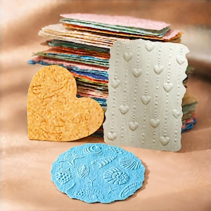 May include: A stack of multicolored paper sheets with rough edges, accompanied by a heart-shaped orange textured paper, a rectangular silver paper with embossed hearts and beads, and a circular blue paper with embossed seashells and starfish.