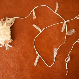 May include: A spool of white lace trim with a length of white lace trim with fringe hanging from it. The lace trim is on a brown surface. The letter H is visible in the lower right corner of the image.