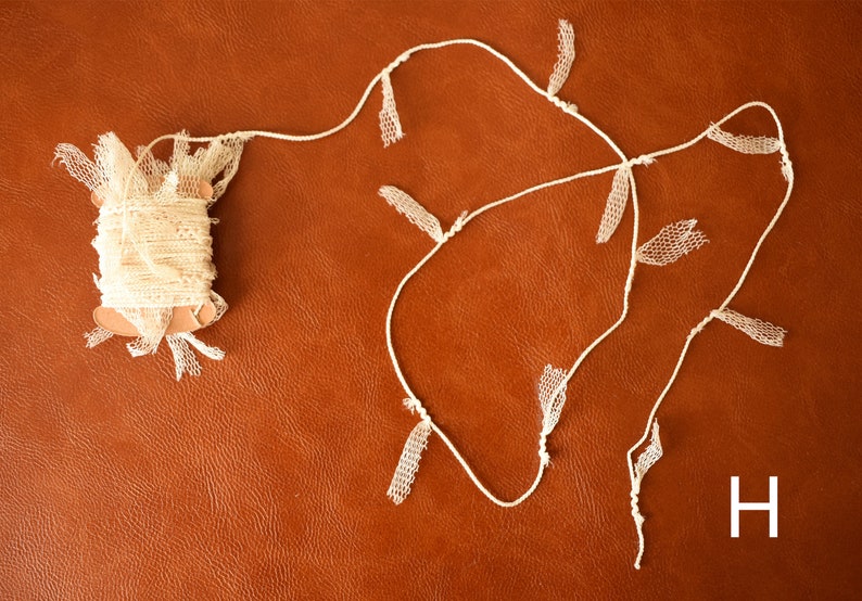 May include: A spool of white lace trim with a length of white lace trim with fringe hanging from it. The lace trim is on a brown surface. The letter H is visible in the lower right corner of the image.