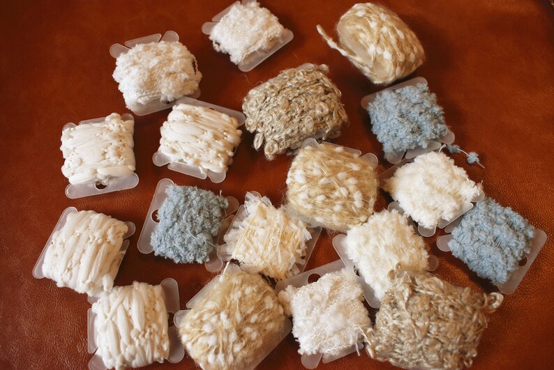 May include: Assortment of white, beige, and blue yarn spools with various textures, including fuzzy, fluffy, and beaded.