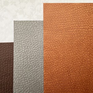 May include: An assortment of textured leather samples in shades of brown, gray, and tan. The leather has a pebbled grain and is arranged in a layered composition, with a floral patterned paper in the background.