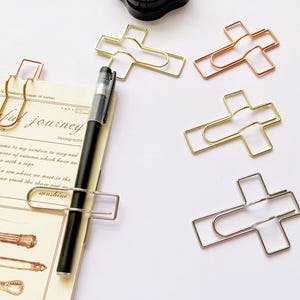 May include: Five gold cross-shaped paper clips. The clips are in different shades of gold, including rose gold. The clips are on a white surface.