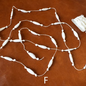 May include: White beaded garland with a spool of white bead garland. The garland is made of small, oval-shaped beads strung on a white thread. The spool is in a clear plastic package. The letter "F" is printed on the brown surface below the garland.
