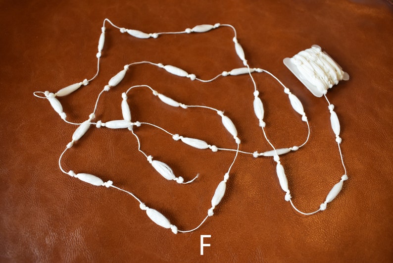 May include: White beaded garland with a spool of white bead garland. The garland is made of small, oval-shaped beads strung on a white thread. The spool is in a clear plastic package. The letter "F" is printed on the brown surface below the garland.