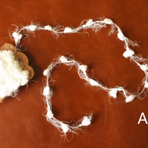May include: A white fluffy garland with small pom poms. The garland is on a brown surface. The letter A is written on the surface.