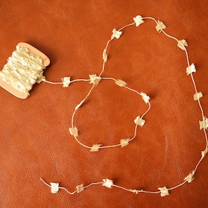 May include: A spool of gold-colored fabric trim with a white background, and a long strand of the trim laid out in a circle on a brown leather surface. The trim is made of small, gold-colored squares.