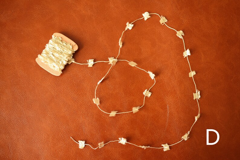May include: A spool of gold-colored fabric trim with a white background, and a long strand of the trim laid out in a circle on a brown leather surface. The trim is made of small, gold-colored squares.