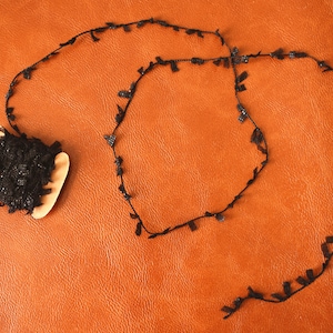 May include: A spool of black lace trim with a decorative fringe. The lace is rolled up on a wooden spool. The lace is on a brown leather surface. The letter "E" is written on the surface.