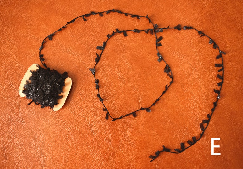 May include: A spool of black lace trim with a decorative fringe. The lace is rolled up on a wooden spool. The lace is on a brown leather surface. The letter "E" is written on the surface.