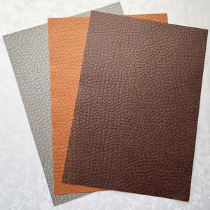 May include: Three sheets of textured faux leather in shades of gray, orange, and brown. The sheets are arranged in a fan shape, showcasing the detailed grain of the material. The colors and texture suggest a craft or design product.