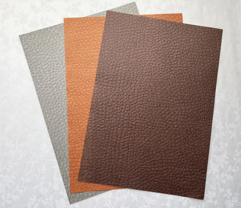 May include: Three sheets of textured faux leather in shades of gray, orange, and brown. The sheets are arranged in a fan shape, showcasing the detailed grain of the material. The colors and texture suggest a craft or design product.