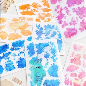 May include: A collection of rectangular paper sheets featuring watercolor-style splatters in shades of orange, pink, and blue. One sheet displays a black circular postmark stamp and the handwritten name Timothy in black ink over blue paint.