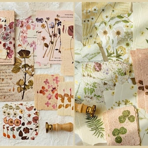 May include: A collection of handmade paper with pressed flowers and botanical designs. The papers feature various floral arrangements in shades of pink, purple, and green. Some papers have text and are accompanied by wooden and brass stamps.