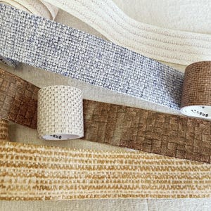 May include: An assortment of decorative tapes in various colors and textures. The tapes include woven patterns in beige, brown, and blue, along with a cream-colored tape with a stitched design. Several rolls of tape are also visible.