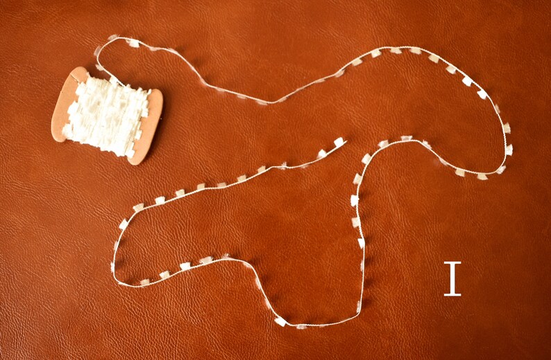 May include: A spool of white trim with a brown cardboard spool. The trim is laid out in a curved shape on a brown leather surface. The letter "I" is written in white on the surface.