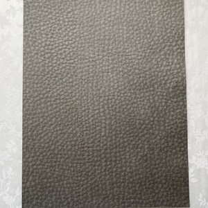 May include: A rectangular sheet of gray faux leather with a textured surface. The word "Gray" is written in a script font at the bottom of the sheet. The background is a white floral pattern.