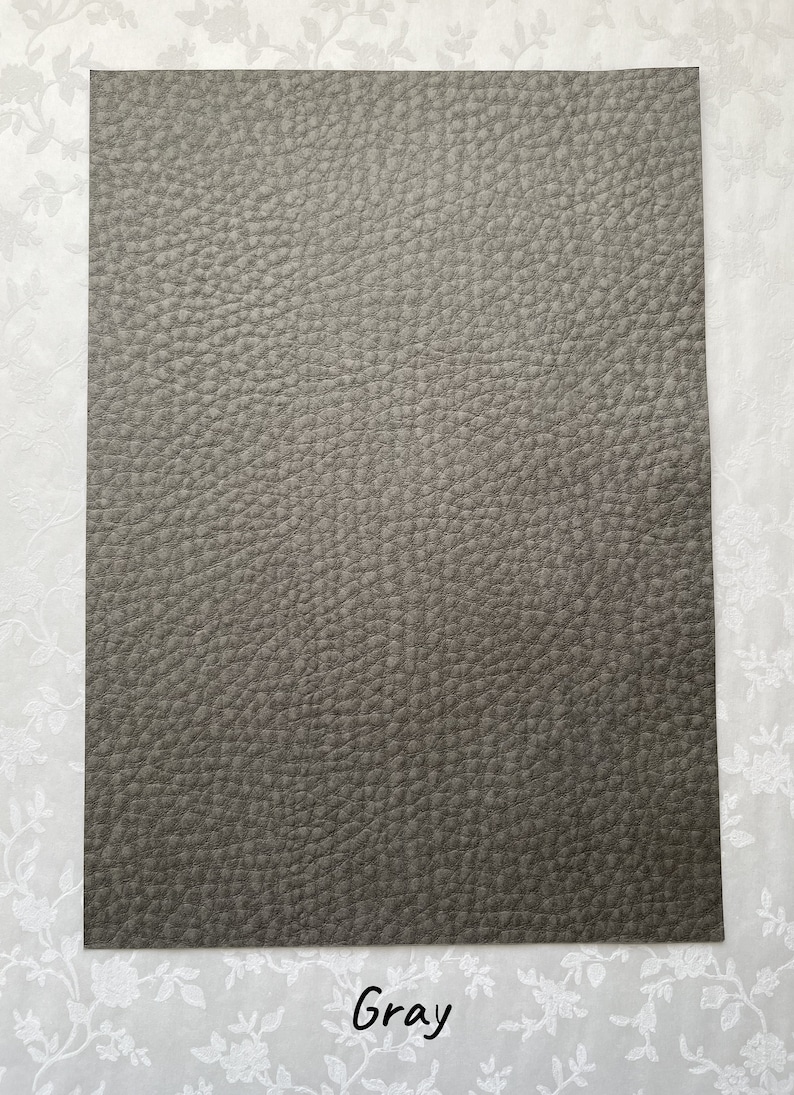 May include: A rectangular sheet of gray faux leather with a textured surface. The word "Gray" is written in a script font at the bottom of the sheet. The background is a white floral pattern.