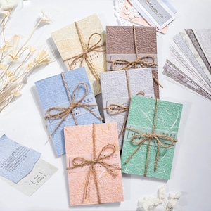 May include: A collection of textured paper notebooks in various pastel colors, tied with twine bows. Each notebook has a small label and is surrounded by additional decorative paper elements. The overall aesthetic is soft and artisanal.