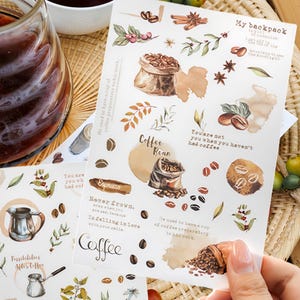 Op de afbeelding: Een set van drie vellen stickers met illustraties en citaten met een koffie-thema. De stickers zijn gedrukt op wit papier met een aquarel-achtige stijl. De stickers bevatten afbeeldingen van koffiebonen, koffiekopjes en koffiezakken. De citaten zijn: "Hij dronk altijd een kopje koffie voordat hij aan het werk ging", "Je bent niet jezelf als je geen koffie hebt gedronken", "Nooit fronsen, zelfs niet als je verdrietig bent, want verliefd worden op je glimlach.", "Koffieboon", "Mijn rugzak", "Mogelijkheden na de koffie", "Anders drinken", en "Koffie".
