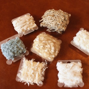 May include: Six spools of yarn in various colors and textures, including white, beige, light blue, and a natural fiber. The spools are arranged on a brown leather surface.
