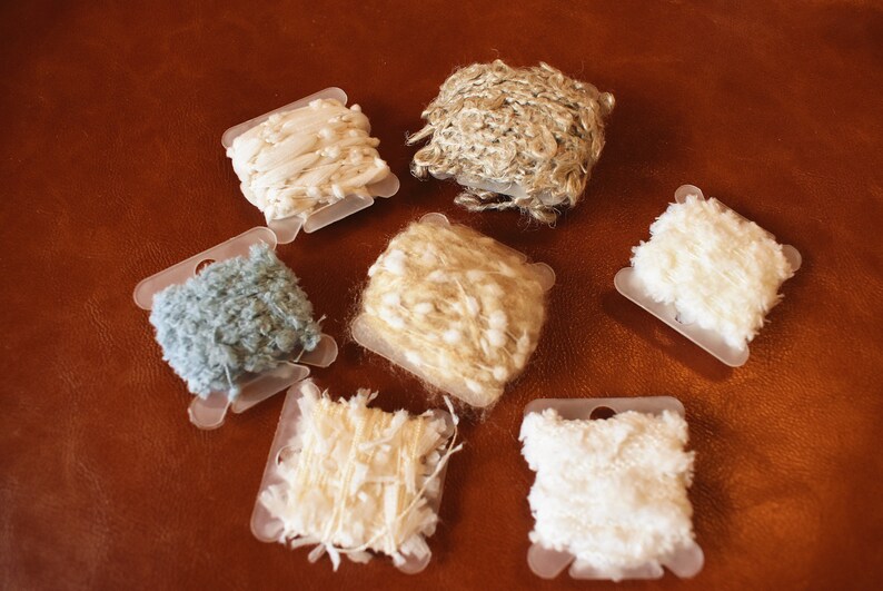 May include: Six spools of yarn in various colors and textures, including white, beige, light blue, and a natural fiber. The spools are arranged on a brown leather surface.