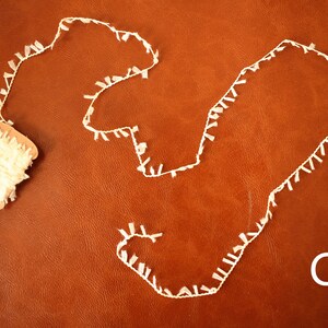May include: A spool of white fringe trim with a length of the trim laid out on a brown surface. The letter "C" is visible in the lower right corner of the image.