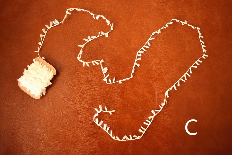 May include: A spool of white fringe trim with a length of the trim laid out on a brown surface. The letter "C" is visible in the lower right corner of the image.