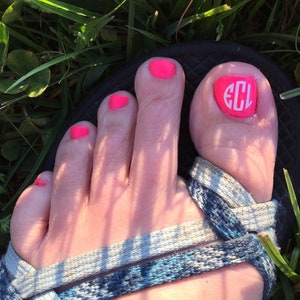 May include: A foot with pink nail polish and a white monogrammed nail sticker that says "ECL" on the big toe.