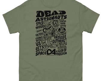 May include: A green t-shirt with a black graphic print of an astronaut in a space suit. The text on the shirt reads "Dead Astronauts" and includes phrases like "Space Invaders", "Come With Us", "Let's Go To Space", and "A Rush of Freedom".