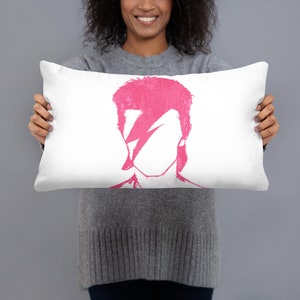 May include: A white pillow with a pink silhouette of David Bowie's face. The silhouette is a simple outline of his face with a lightning bolt over his eye.