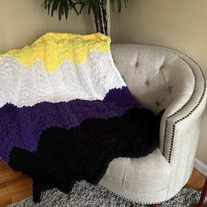 May include: A crocheted blanket with yellow, white, purple, and black stripes. The blanket is draped over a light gray armchair.
