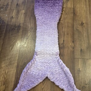 May include: A purple and white ombre mermaid tail blanket made of soft, fluffy yarn. The tail is crocheted and has a textured, scaly appearance.