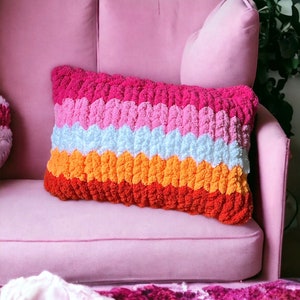 May include: A multi-colored knitted throw pillow with a pink, white, orange, and red striped pattern. The pillow is sitting on a pink couch.