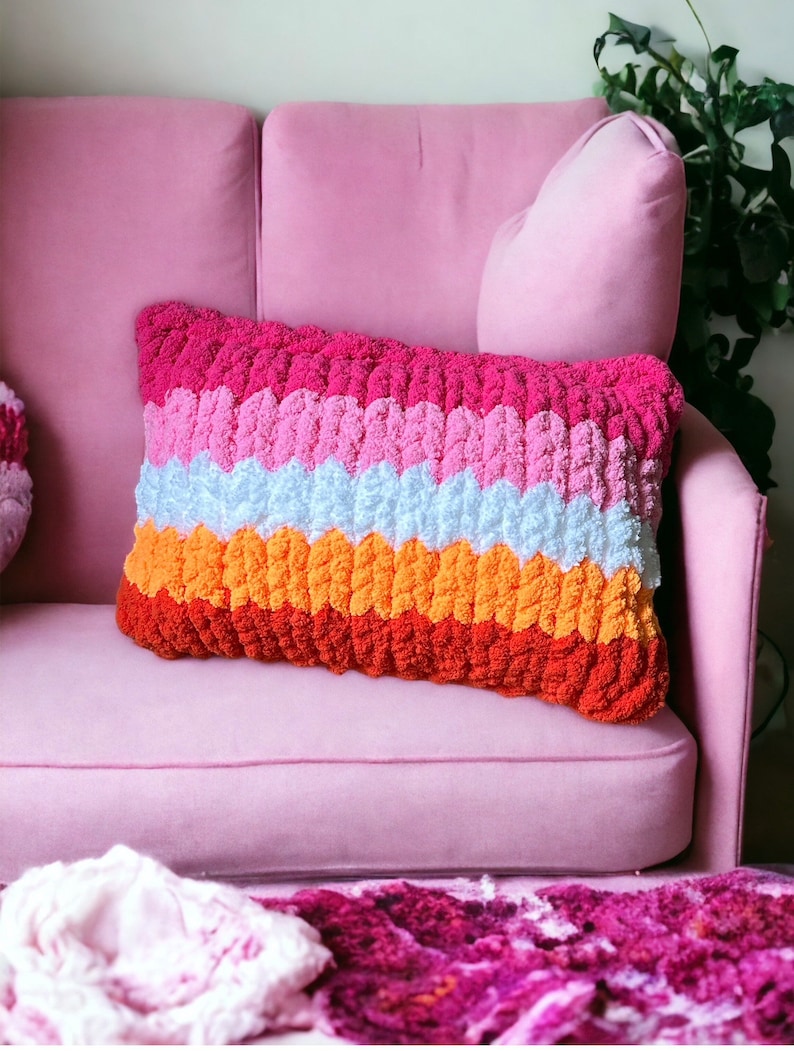 May include: A multi-colored knitted throw pillow with a pink, white, orange, and red striped pattern. The pillow is sitting on a pink couch.