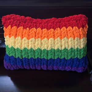 Puede incluir: Una almohada de crochet de colores arco&iacute;ris con una textura esponjosa. La almohada est&aacute; hecha con diferentes tonos de hilo rojo, naranja, amarillo, verde, azul y morado.