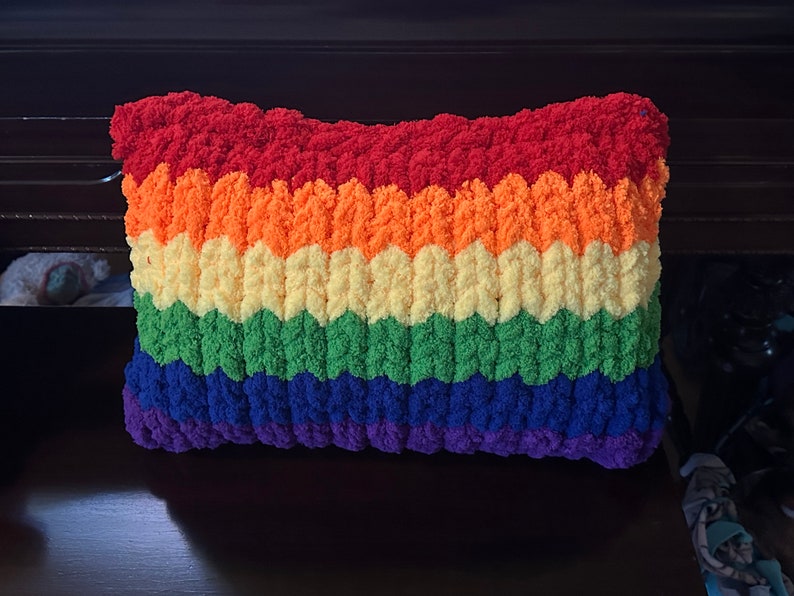 Puede incluir: Una almohada de crochet de colores arco&iacute;ris con una textura esponjosa. La almohada est&aacute; hecha con diferentes tonos de hilo rojo, naranja, amarillo, verde, azul y morado.