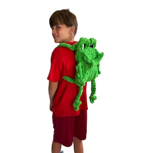 May include: A bright green crochet backpack shaped like a frog, featuring a textured yarn surface, two shoulder straps, and dangling limbs. The frog design includes prominent eyes and a wide mouth, crafted from soft, chunky green yarn material.