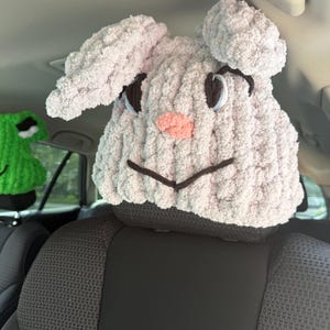 May include: A light grey crocheted headrest cover shaped like a rabbit with long floppy ears, dark brown embroidered eyes, a small pink nose, and a stitched mouth. The plush, textured yarn creates a soft appearance, fitting snugly over a car seat headrest.