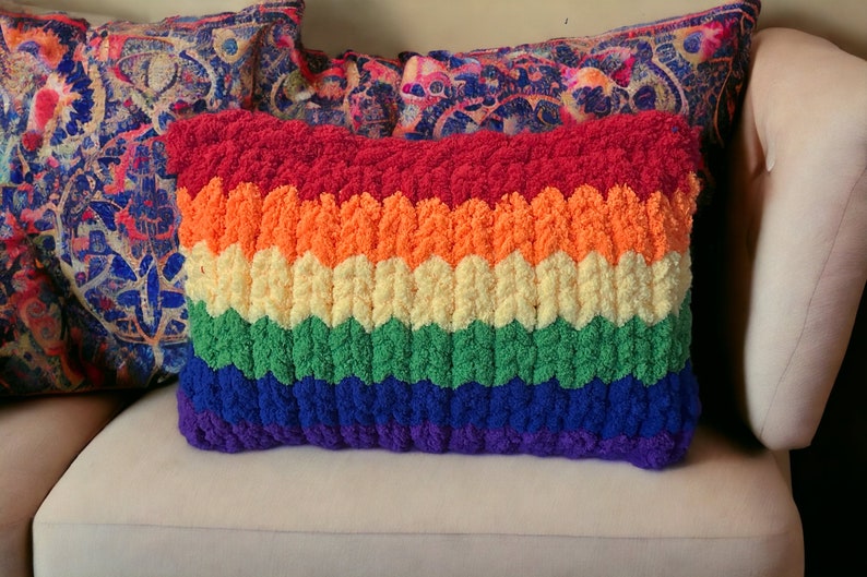 Puede incluir: Una almohada de crochet de colores arco&iacute;ris con una superficie texturizada. La almohada est&aacute; hecha de hilo rojo, naranja, amarillo, verde, azul y morado.