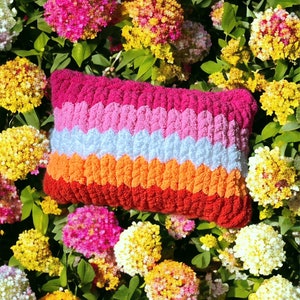 May include: A colorful striped pillow with a textured, crocheted design. The pillow is made of pink, white, orange, and red yarn. The pillow is lying on a bed of pink, white, and yellow flowers.