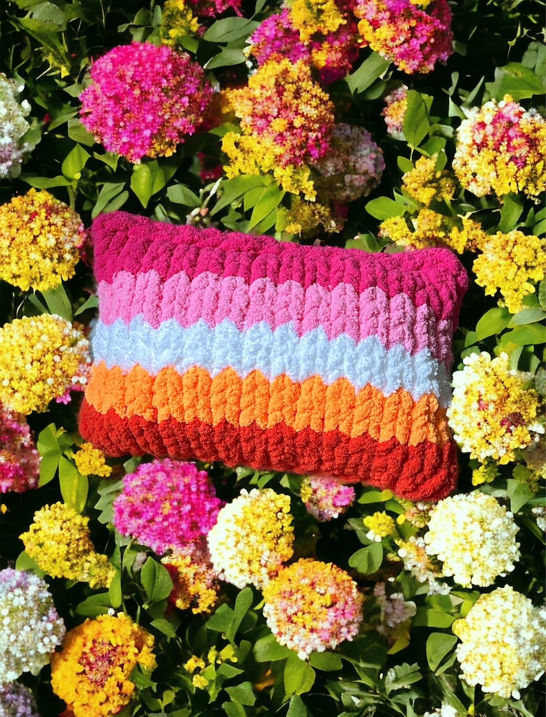 May include: A colorful striped pillow with a textured, crocheted design. The pillow is made of pink, white, orange, and red yarn. The pillow is lying on a bed of pink, white, and yellow flowers.