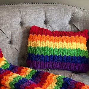 Puede incluir: Una manta y una almohada de crochet a rayas de arco&iacute;ris en un sof&aacute; gris. La manta y la almohada est&aacute;n hechas con hilo grueso y tienen una textura esponjosa.