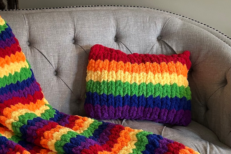 Puede incluir: Una manta y una almohada de crochet a rayas de arco&iacute;ris en un sof&aacute; gris. La manta y la almohada est&aacute;n hechas con hilo grueso y tienen una textura esponjosa.