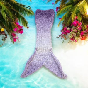 May include: A purple and white mermaid tail blanket with a fluffy texture. The blanket is lying on a blue and white background that resembles a beach.