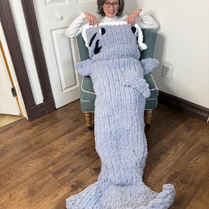 May include: A grey knitted shark blanket with a white and black face. The blanket is large enough to cover a person sitting in a chair.