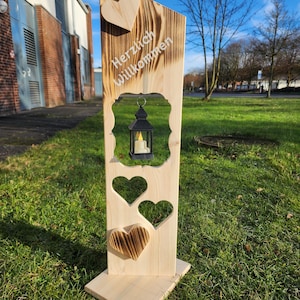 Könnte beinhalten: Ein hölzernes Willkommensschild mit einem Herz-Ausschnitt und den Worten "Herzlich Willkommen" in weißer Schrift. Das Schild hat eine kleine Laterne mit einer Kerze darin und drei Herz-Ausschnitte unten.