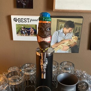 May include: A black and silver beer tap with a colorful tiki head spout. The tap is surrounded by glasses and a silver stein with a crest. The tap is on a black counter with a silver metal mat. There is a silver garland on the counter and a brown leather chair in the background.