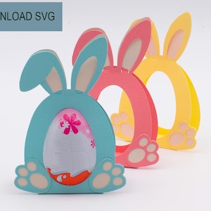 Op de afbeelding: Drie kleurrijke paashaasvormige decoraties. De eerste is lichtblauw met een wit en roze ei erin. De tweede is roze en de derde is geel. De tekst "DIGITAL DOWNLOAD SVG" staat linksboven.