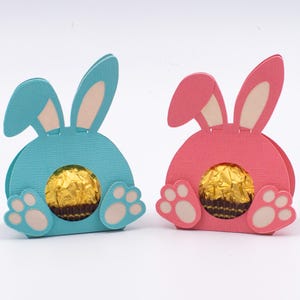 May include: Two Easter-themed paper bunny treat holders. One is light blue, the other is pink. Each has a round opening with a gold-wrapped chocolate candy inside. The bunny's ears and feet are visible.